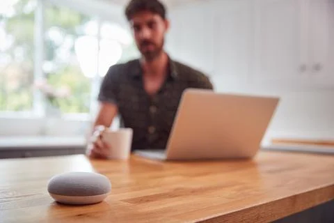 Man In Kitchen Using Digital Smart Speaker Whilst Working From Home With Laptop 스톡 사진