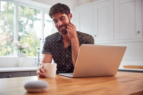 Man In Kitchen Using Digital Smart Speaker Whilst Working From Home With Laptop 스톡 사진