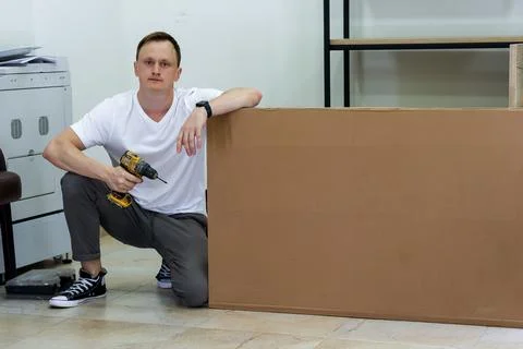 Man kneeling with drill leaning on large cardboard box in office environment Stock Photos