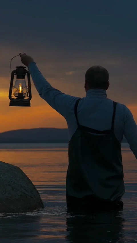 A man with a lantern in his hands stands on the shore of a lake at sunset. Video stock 336620962