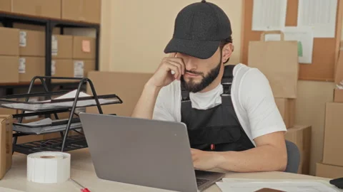 Man at laptop among parcels, hand to temple rubbing eyes at packing desk in.. Stock Footage 337371840