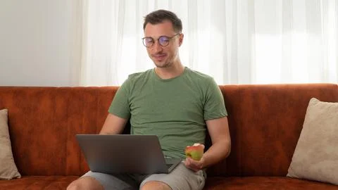 A man with a laptop on the couch eats an apple Stock Photos