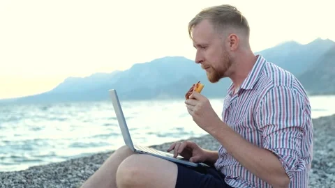 Man with laptop eating pizza while sitting on the beach 스톡 동영상 220865882