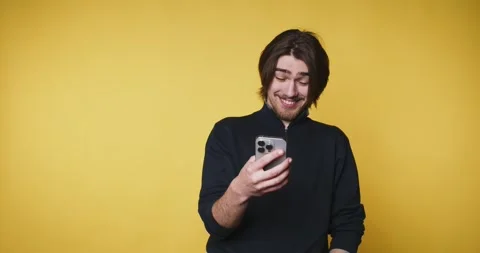 Man laughs while reading messages on his phone against a yellow background .. Видео 328249953