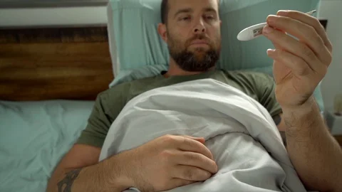 Man laying in bed being sick checking his temperature Видео 128294411