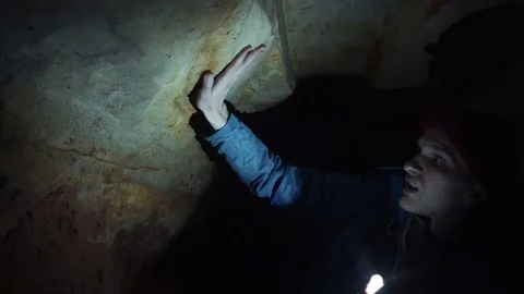 Man laying on ground into narrow cave explores cave walls with hand flashlight Stock Footage 80053577