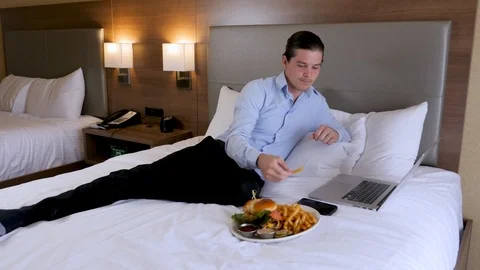 Man lays on hotel bed while working and eating Stockbeeldmateriaal 94267352