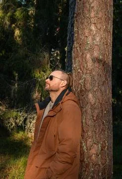 A man leaning against a tree in a forest, wearing sunglasses and a brown jacket Foto stock