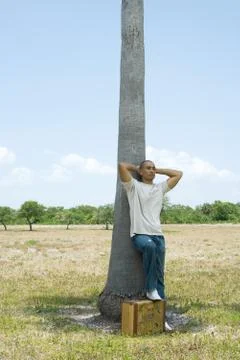 Man leaning against tree trunk, hands behind head, one foot resting on suitcase Stock Photos