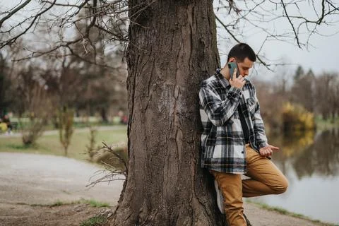 Man leans against tree while talking on phone in a calm park setting Stock Photos