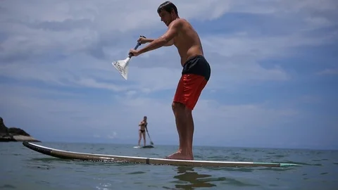 A man learns to stand on a longboard, can not hold the balance with a paddle in Stock Footage 95937535