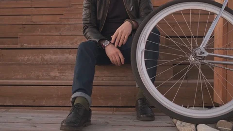 A man in a leather jacket and jeans sits on the wooden steps near the bike and Stockbeeldmateriaal 121761202