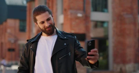 Man in leather jacket standing in street, using smartphone, makes selfie on it. Stock Footage 246753857