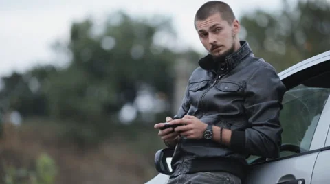 The man in the leather jacket is typing a message near the car Stock Footage 55334069