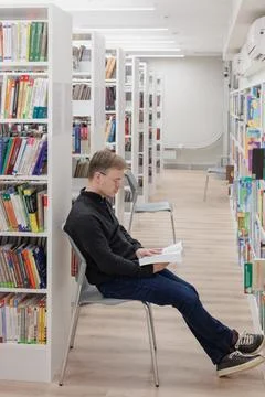 Man in library is reading book sitting on chair near bookshelves. Stock Photos