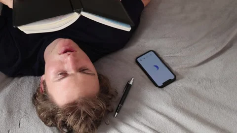 Man lies on bed with closed eyes beside smartphone glowing on blanket and pen ne Stock Footage 314979736