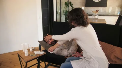 Man lies in bed feeling sick, sneezing and coughing, young girl doctor helps to Stock Footage 201877309