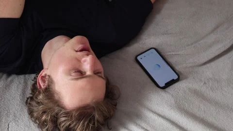 Man lies on bed next to smartphone with glowing screen speaking thoughtfully atm Video stock 314979714