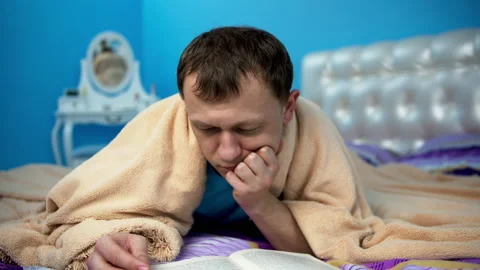 A man lies on a bed under a blanket and reads a book Stock Footage 132052265