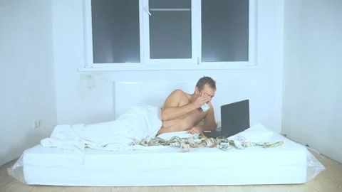 A man lies on a mattress in room, uses a laptop, wipes his tears with cash Stock Footage 143695053