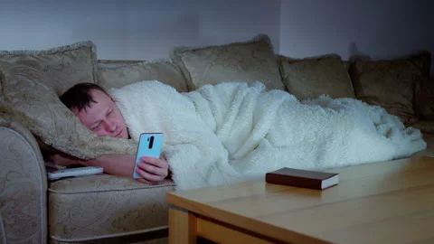 A man lies on the sofa in front of the television at night with his mobile phone Stock Footage 319383430
