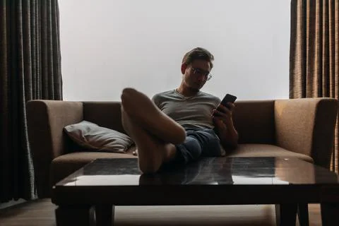 Man lies on sofa using phone with his feet on the table. Concept of manner. Stock Photos