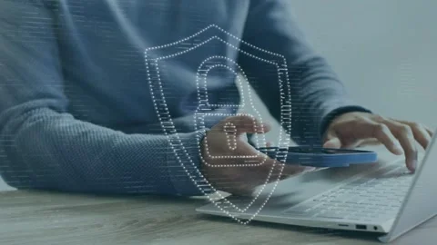 Man lifting blue phone, tapping laptop keys at desk, forming shield over devices Stock Footage 335761985