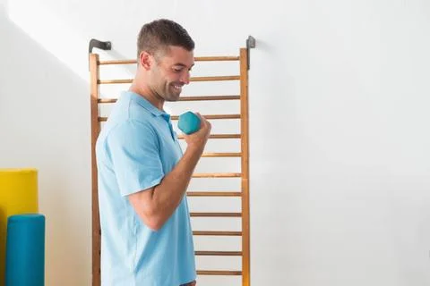 Man lifting hex dumbbell by stall bars with foam rollers in rehab room wearing Stock Photos