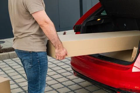 Man lifting long cardboard box from trunk of red sedan during home delivery Stock Photos