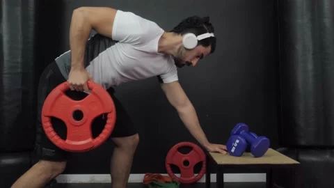 Man lifting red olympic weight disc with one hand indoors while listening Video stock 151854389