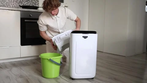 Man in light clothes empties full water tank from home dehumidifier into green b Stock Footage 318867822