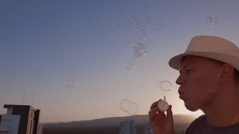 A man in a light-colored hat blowing soap bubbles, against the sky at sunset Stock Footage 307932899