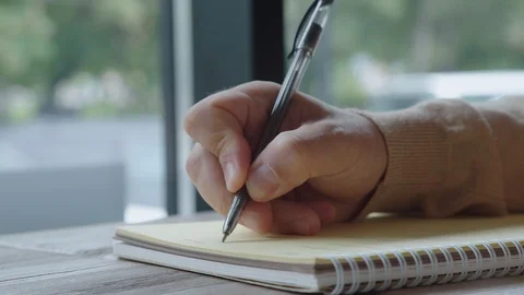 Man in light sweater writes in a notebook with a black pen, close-up. Vidéo 93561080