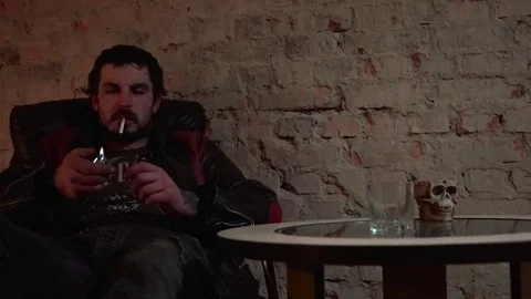 Man lights cigarette and puts the lighter on the table next to glass and skull 스톡 동영상 84184759