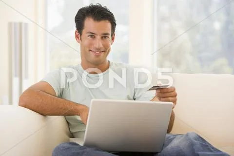 Man in living room using laptop and holding credit card smiling Stock-Fotos