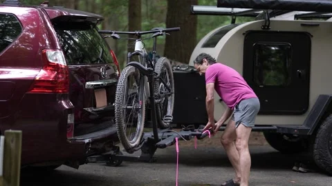 Man Loading Bicycles Onto Car Near Teardrop Trailer at Wooded Campground Stock Footage 313899236