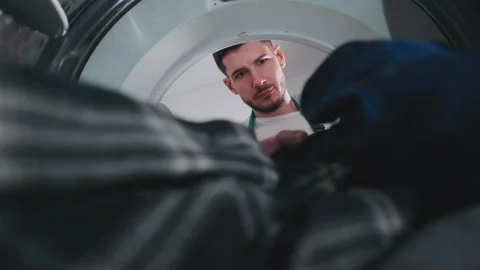 Man loads laundry into a dryer while wearing an apron in a modern laundry room Stock Footage 305280507