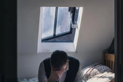 A man looking down with head in hand, wearing a waistcoat sitting on a bed Stock Photos