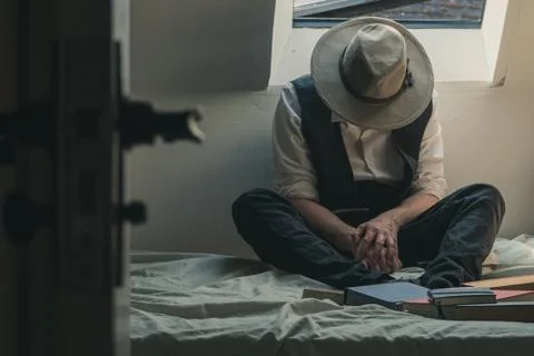A man looking down wearing a fedora hat and waistcoat sitting on a bed Stock Photos