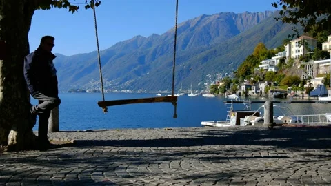 Man Looking at an Empty Swing on the Waterfront with Mountain in a Sunny Day Stock Footage 164895546