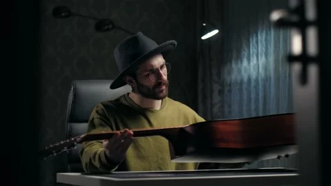 Man looking at a guitar at home.Young guy wear hat repair guitar sitting indoor Stock Footage 232132453