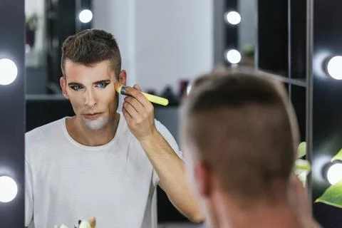 Man looking himself in the mirror while applying makeup with brush. Stock Photos