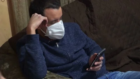 A man looking at his cell phone in the living room wearing a mask Stockbeeldmateriaal 155303481