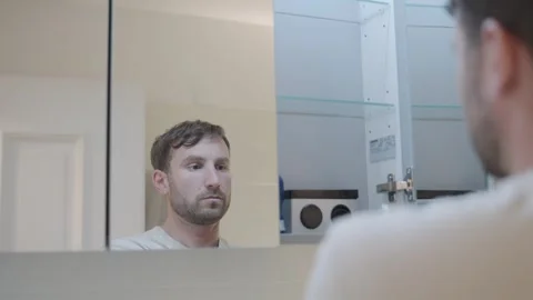 Man looking at his reflection in the bathroom mirror opening a cabinet for Stock-Footage 335374164