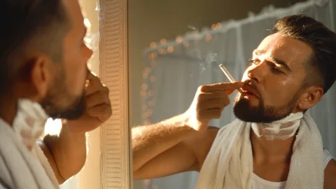 Man  looking in the mirror, smoking a cigarette and shaving his beard 動画素材 116334861
