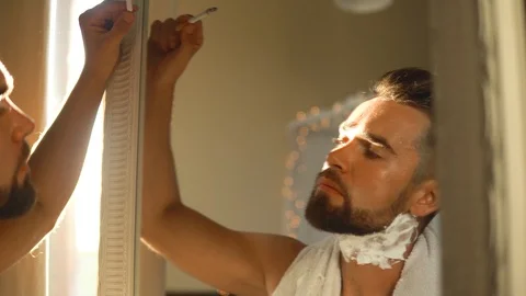 Man  looking in the mirror, smoking a cigarette and shaving his beard Stock-Footage 116334901
