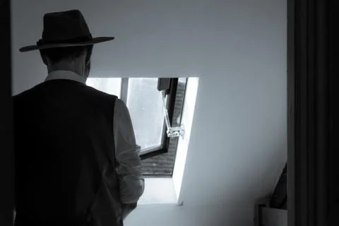 A man looking out of a window, back to camera, wearing a waistcoat and hat. Stock Photos