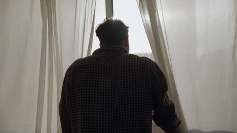 Man looking out window, backshot. peeking his head between white curtains. sun 库存影片 315755519