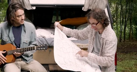 Man looking at a paper map while his male friend plays guitar while camping Stock Footage 80809519