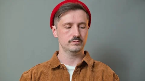 A man, looking sadly down, wearing a red hat and a brown jacket, stands calmly Vidéo 337642640